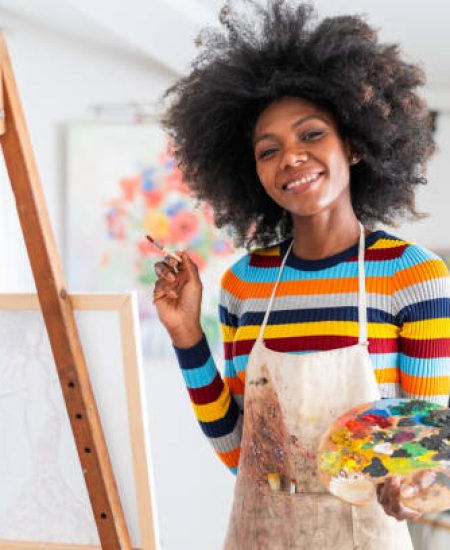 Smiling african female fine artist with afro hair and wearing an apron, drawing in a studio on a canvas, placed on a tripod